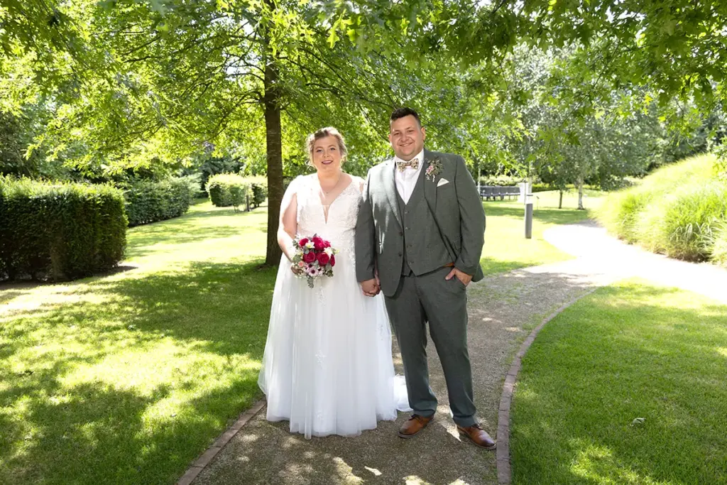 brautpaar steht hand in hand im grünen auf einem Parkweg, natürliche Hochzeitsfotografie im Freien